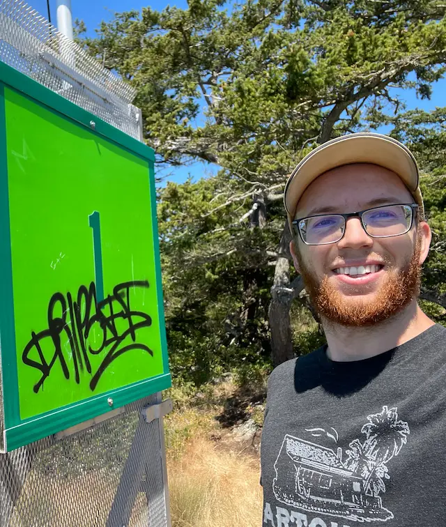 Sam standing next to a beacon with graffiti on it in Lighthouse Point State Park, Washington, smiling as if he was the one who put it there.